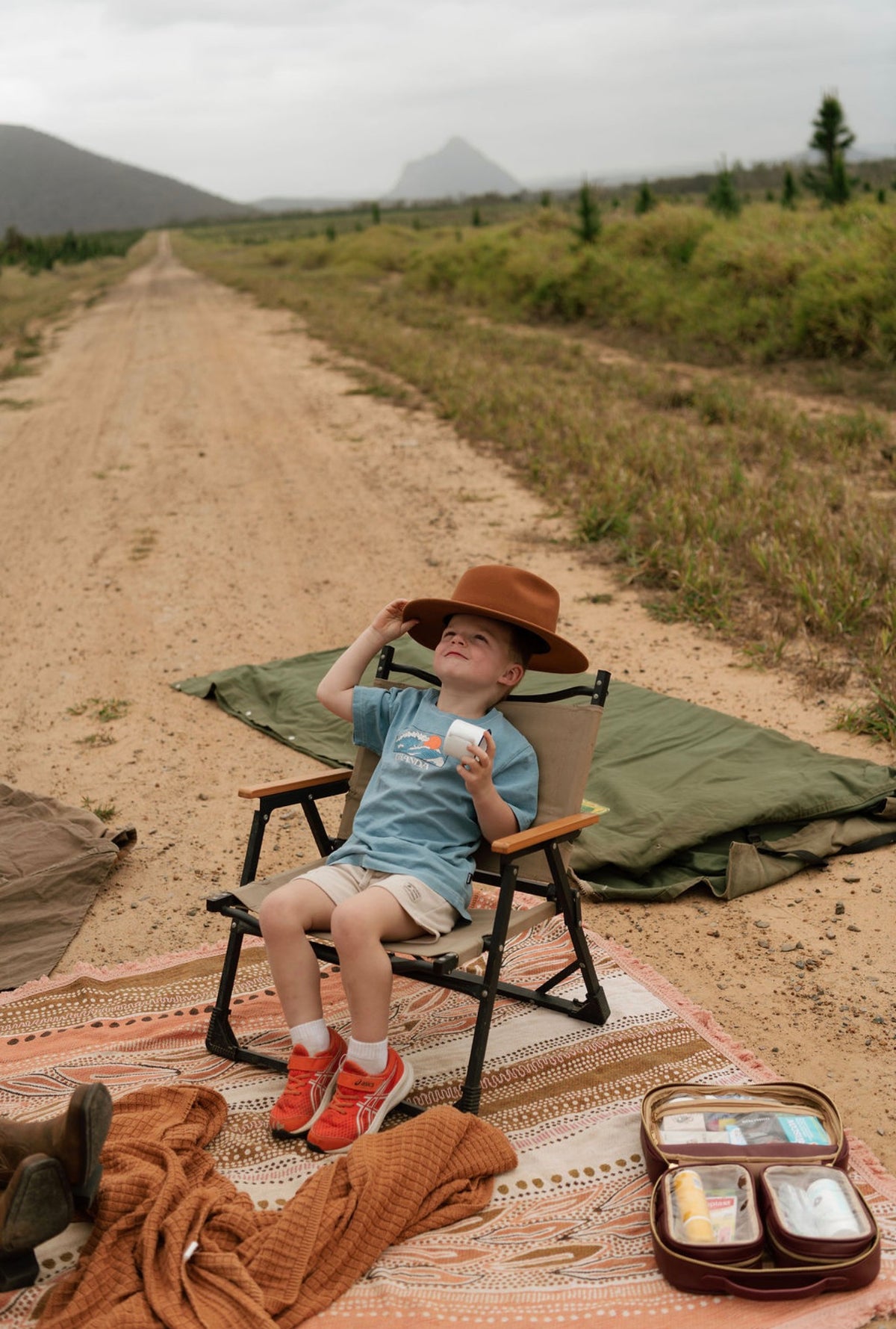 Lifepac Ouchie Kit with first aid supplies, placed on a patterned outdoor rug next to a young boy sitting in a camping chair on a dirt road.